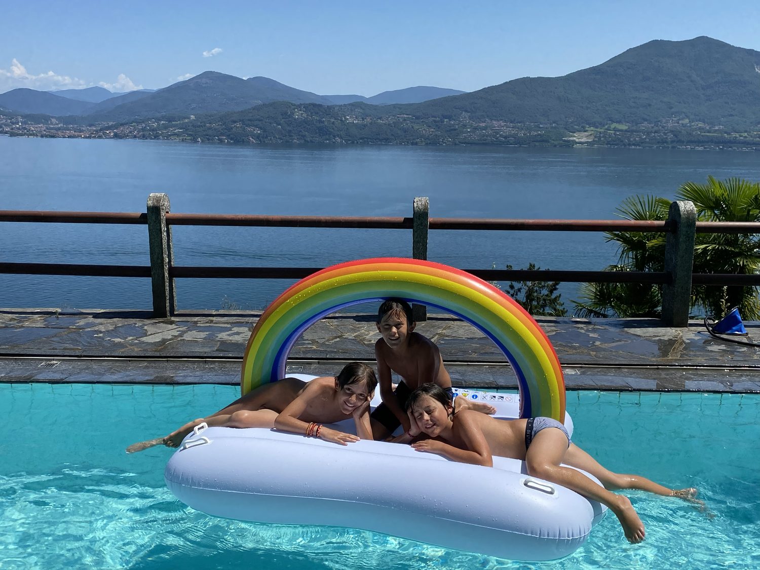 Three children on a rainbow float in the pool, the lake behind